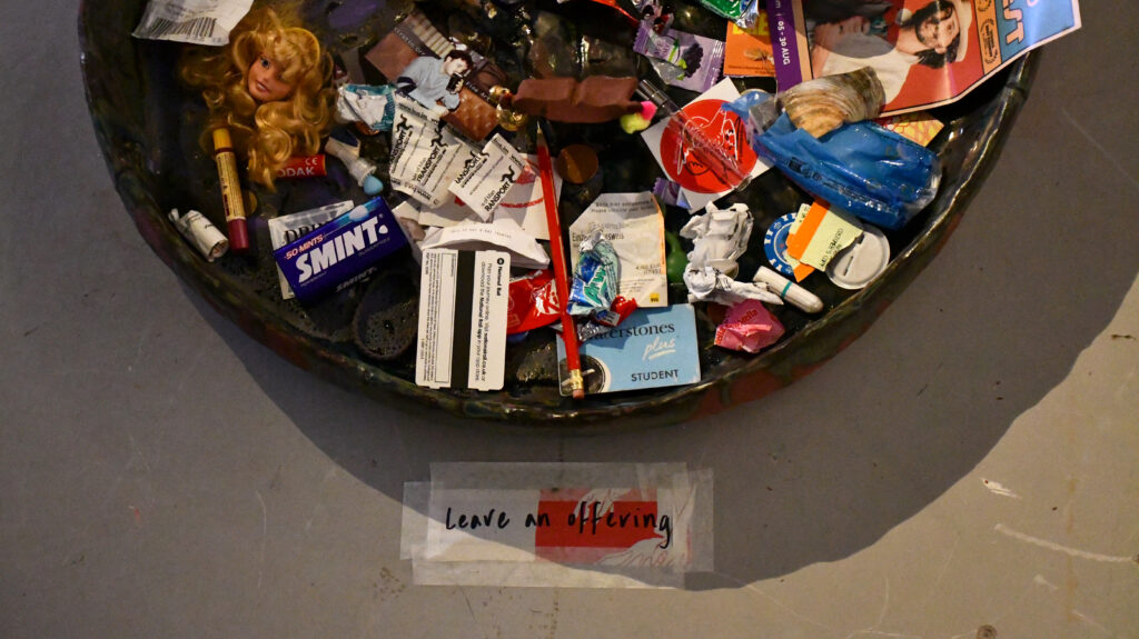 a large bowl holds an array of different objects including train tickets, chewing gum, a barbie head, and old receipts, a sign on the floor next to the bowl reads 'leave on offering'