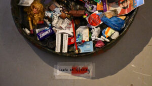 a large bowl holds an array of different objects including train tickets, chewing gum, a barbie head, and old receipts, a sign on the floor next to the bowl reads 'leave on offering'
