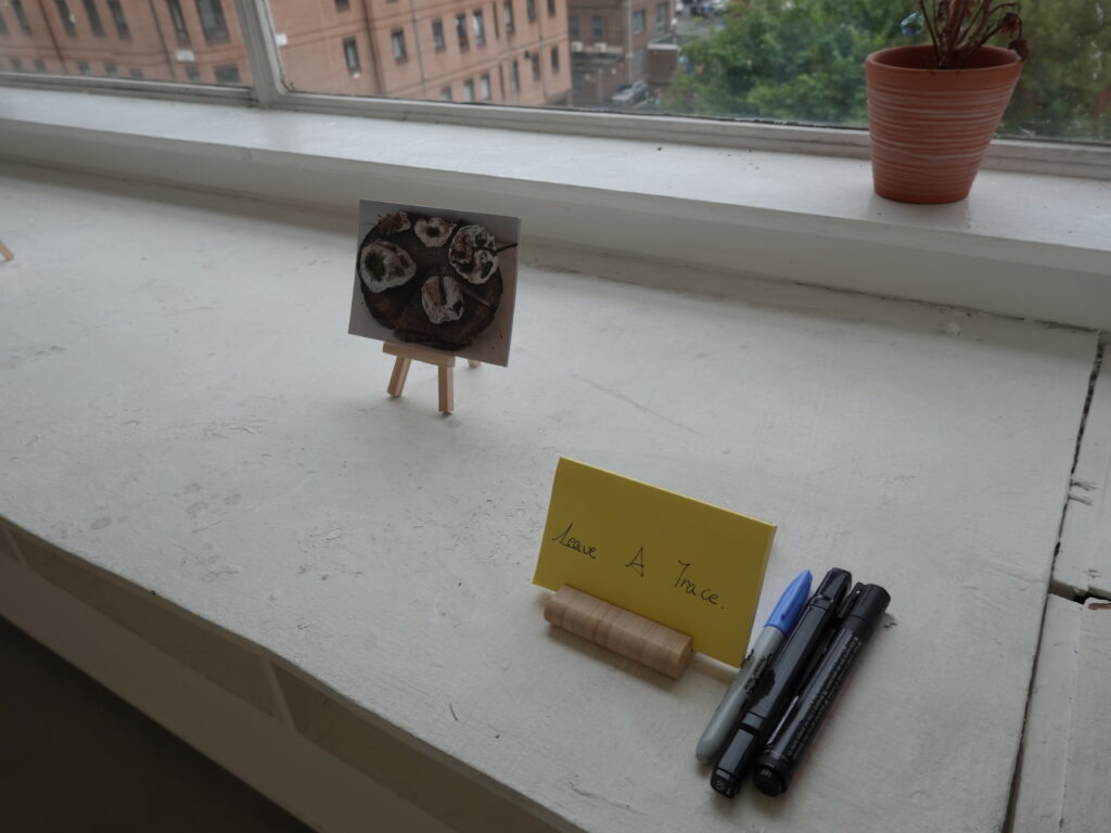 Detail of the Tending the Trace audience response area showing a yellow “Leave A Trace” card, pens, and a small image displayed on a wooden easel.