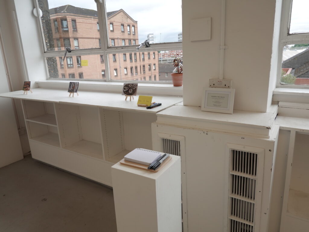 Installation view of the Tending the Trace audience response area, showing small images on wooden easels along a windowsill, a framed project text, and a notebook with pens on a white plinth.