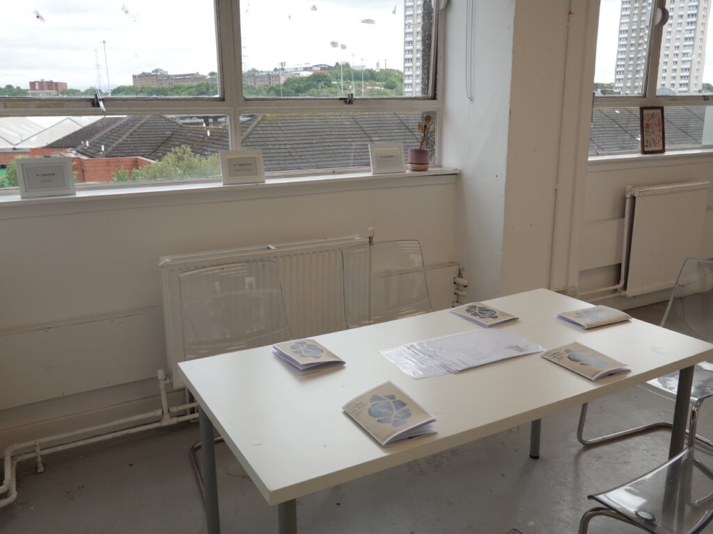 Installation view of the Tending the Trace reading space, showing publications arranged on a white table with framed section texts displayed along the windowsill.