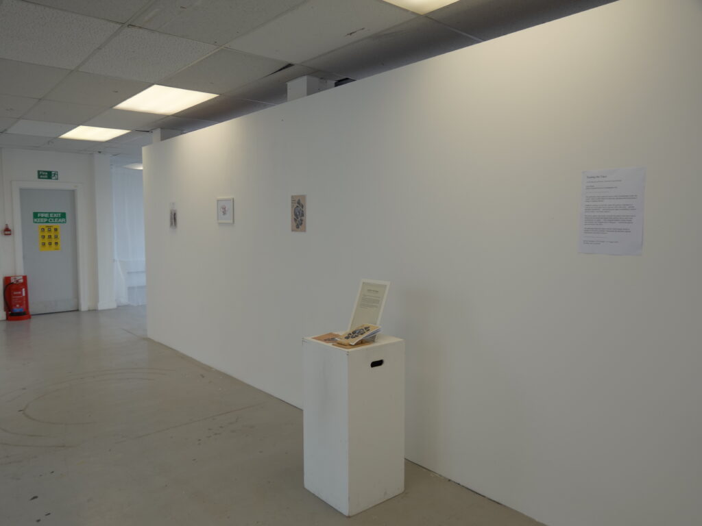 Installation view of Tending the Trace showing a long white exhibition wall, a publication displayed on a white plinth, and framed and printed materials.