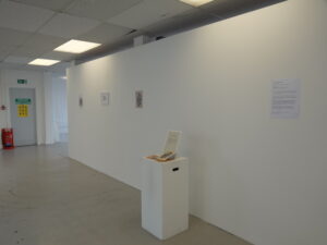 Installation view of Tending the Trace showing a long white exhibition wall, a publication displayed on a white plinth, and framed and printed materials.