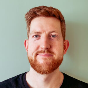 A headshot of a white man in his 30s with short red hair, a short red beard and blue eyes. He is smiling.
