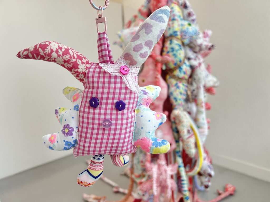 A small handmade textile object held in front of the large suspended ABEBE installation.