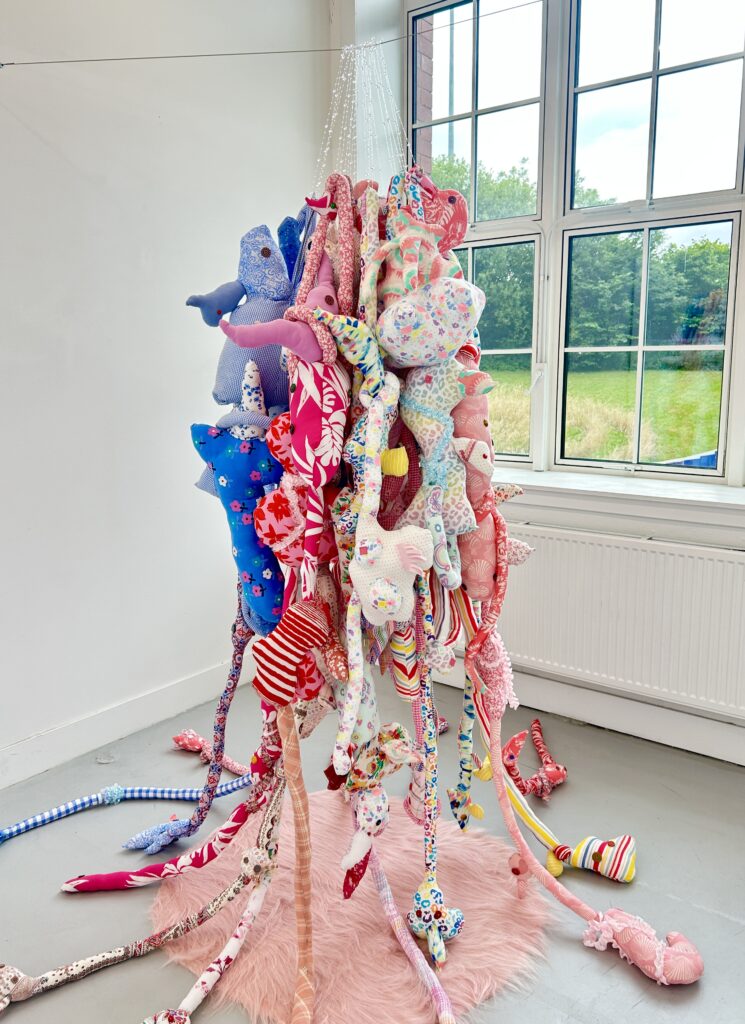 A suspended soft sculpture made from colourful second-hand textiles in a gallery space.