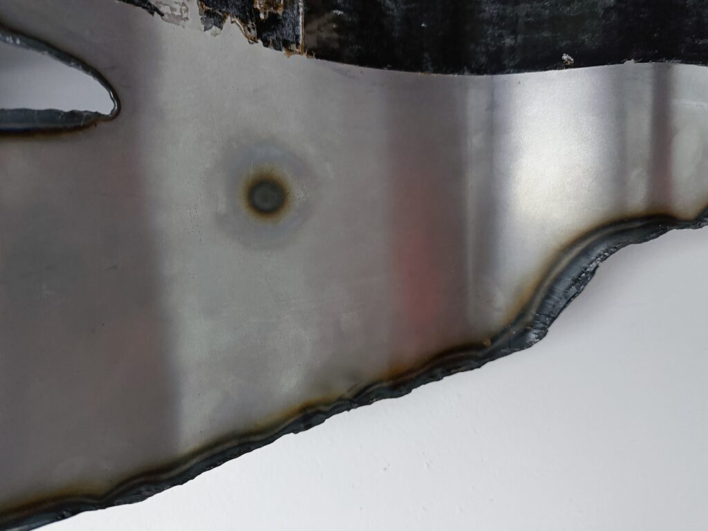 Close-up of a plasma-cut steel edge showing dark heat marks and a circular burn trace on the metal surface.