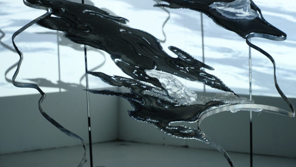 Close-up of plasma-cut steel forms with a transparent resin element, illuminated by projected water imagery.
