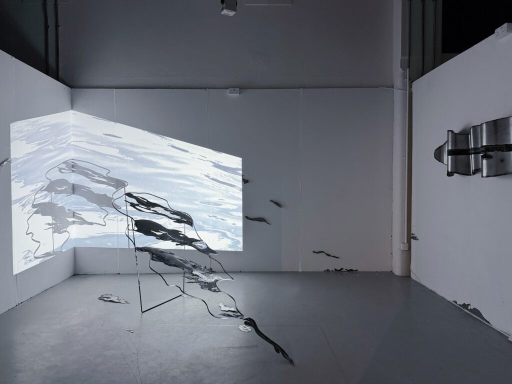 Wide installation view showing a steel sculpture extending across the floor, projected water imagery on the walls, and wall-mounted steel elements.