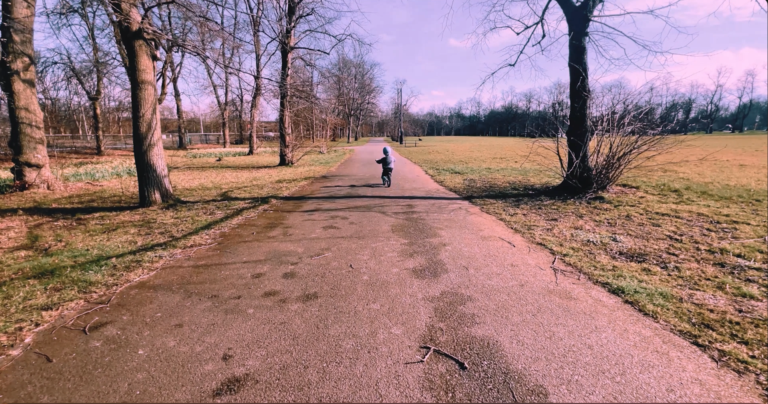 Still from a film showing a wide shot of a path in a park stretching into the distance. Sparse bare trees line the path. In the middle distance a young child is riding a balance bike away from the camera.