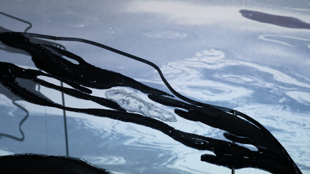 Close-up of dark steel forms and a transparent resin element against projected rippling water imagery.
