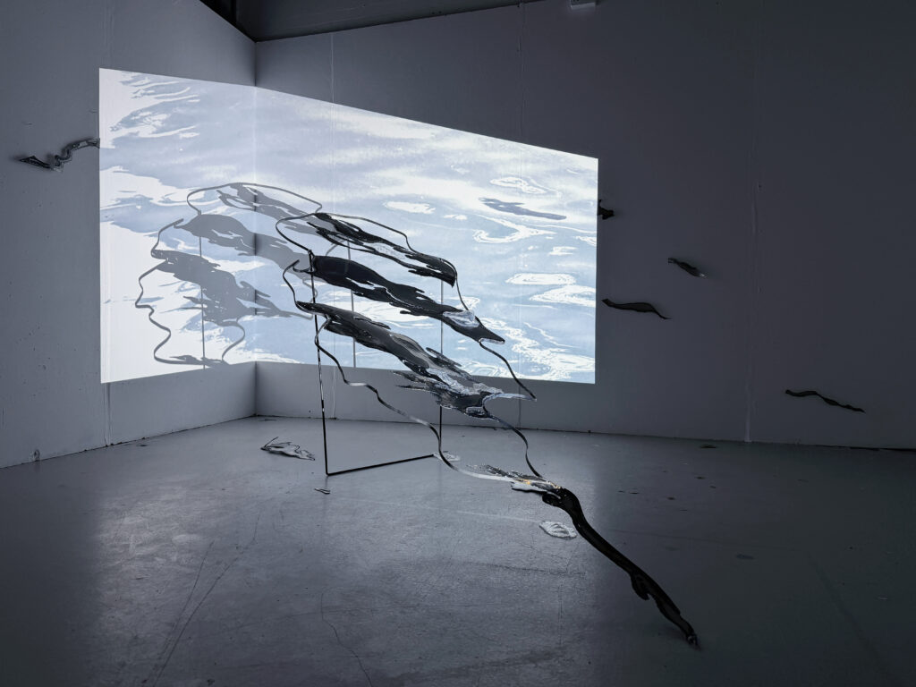 Installation view of a steel sculpture with projected water imagery extending across the wall in a dark exhibition space.