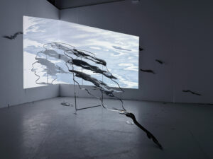 Installation view of a steel sculpture with projected water imagery extending across the wall in a dark exhibition space.