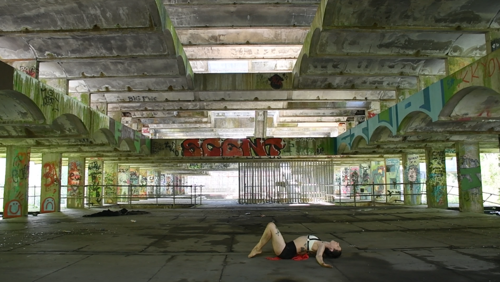 a person wearing black shorts and white latex top lies on the floor of an abandoned concrete building covered in graffiti