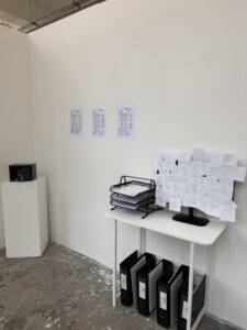 Installation view of a fictional artist agency office with archival folders, documents, handwritten notes and a monitor.