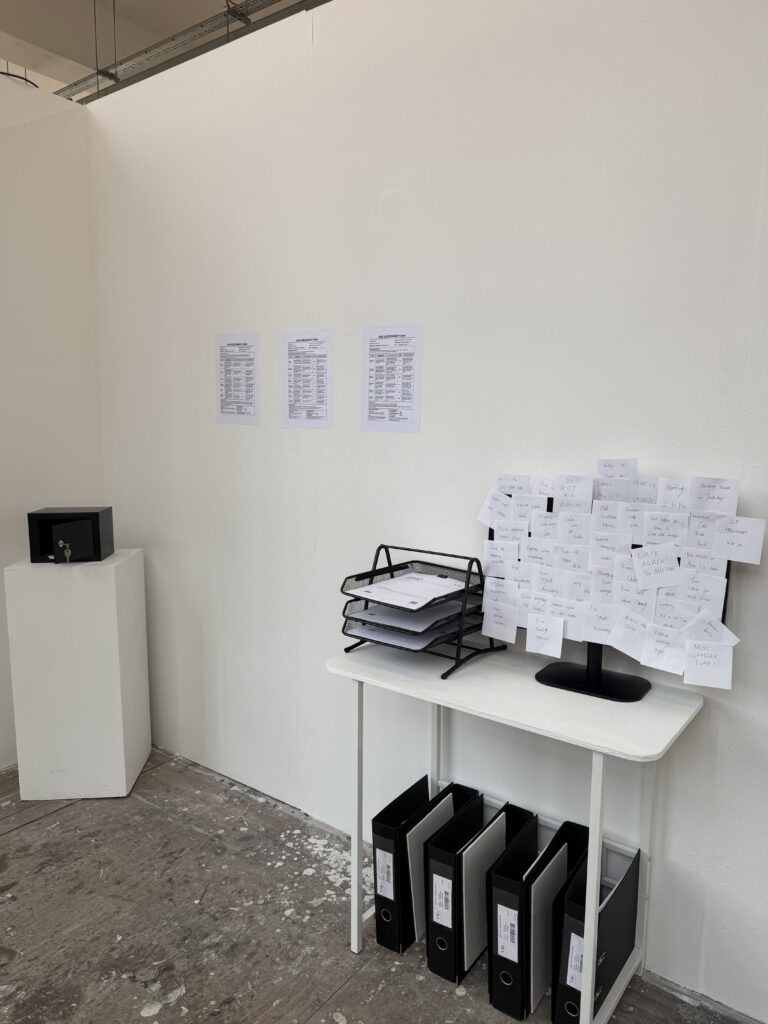 Installation view of a fictional artist agency office with archival folders, documents, handwritten notes and a monitor.