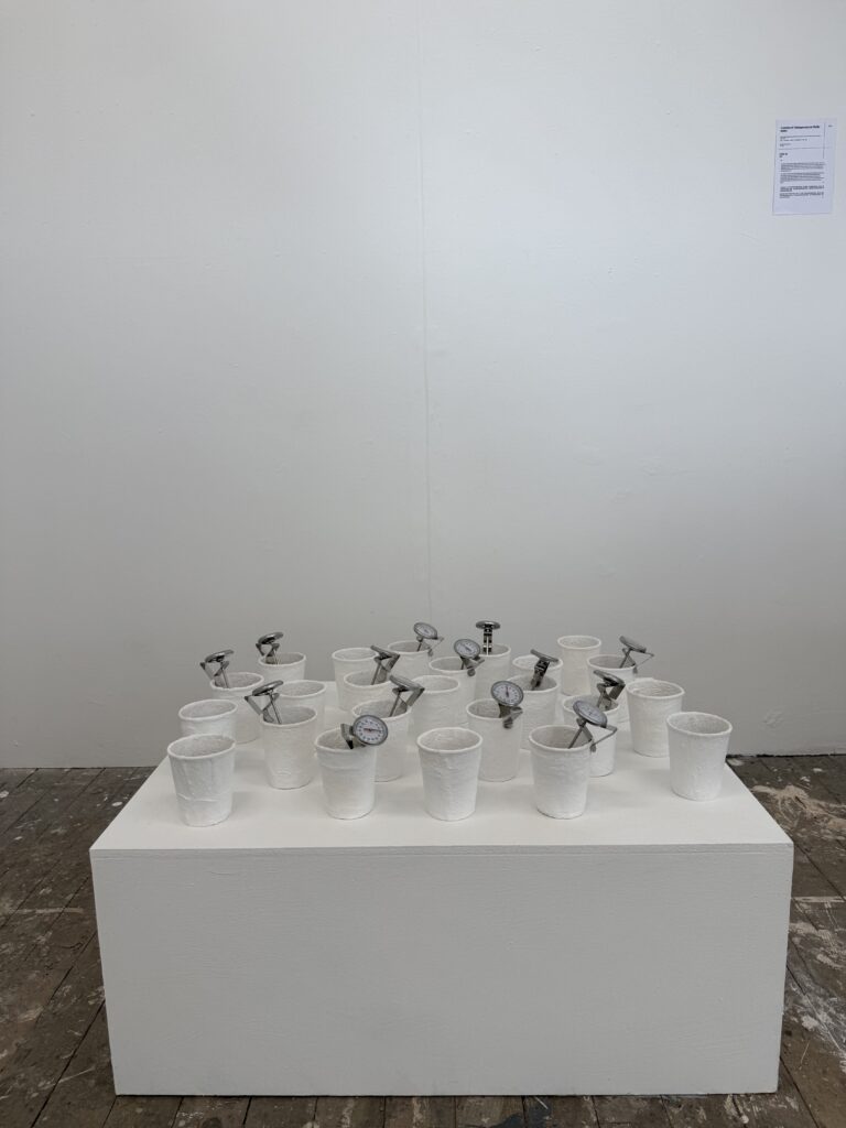 Installation of plaster-wrapped coffee cups and analogue temperature gauges arranged on a low white plinth.