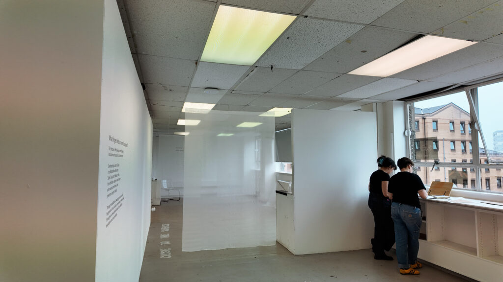 Two visitors viewing materials by the window in The Uncanny: What Remains, with translucent curtains and wall text visible across the exhibition space