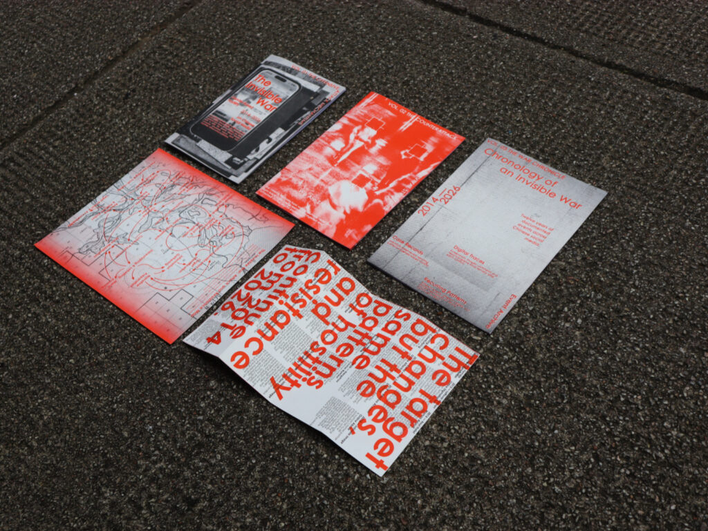 Five red, grey, black and white printed pieces are arranged separately on dark asphalt. Three closed books show a phone-image cover, a red photographic cover and a grey chronological archive cover. A map print lies on the left, and an open publication with oversized red text lies in the foreground.