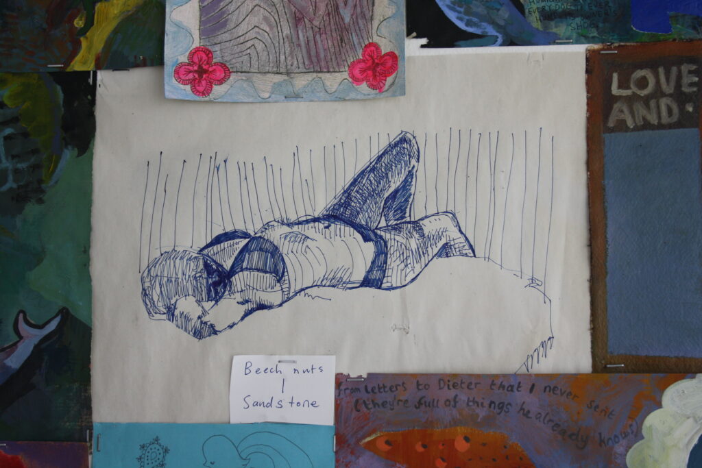 A close up shot of multiple drawings and paintings tacked to the wall. In the centre is a drawing of a model completed in blue pen.