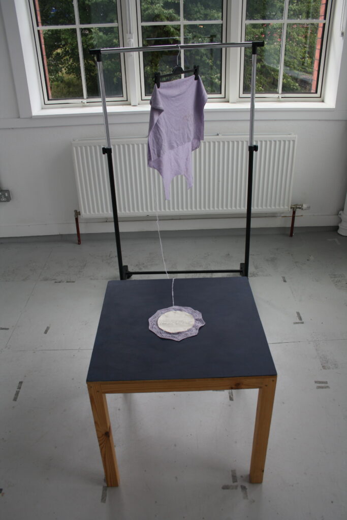 A small knit lace doily on a dark blue table. Behind it is the partially-unravelled back panel of a jumper hanging on a clothing rail. The doily is knit from the same yarn unravelled from the jumper.