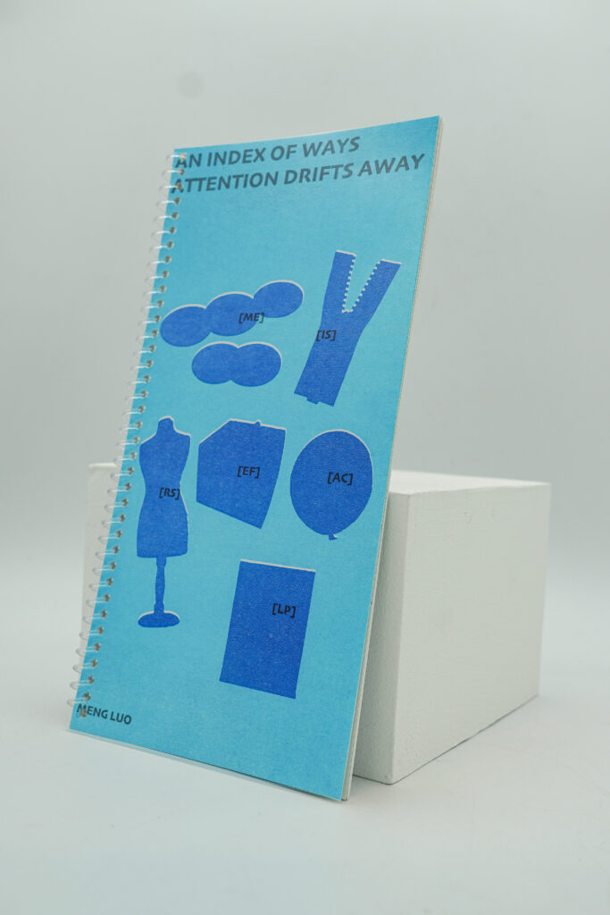 A blue spiral-bound publication titled An Index of Ways Attention Drifts Away stands upright against a white display block. The cover features six blue graphic symbols representing the project's attention-drift families.