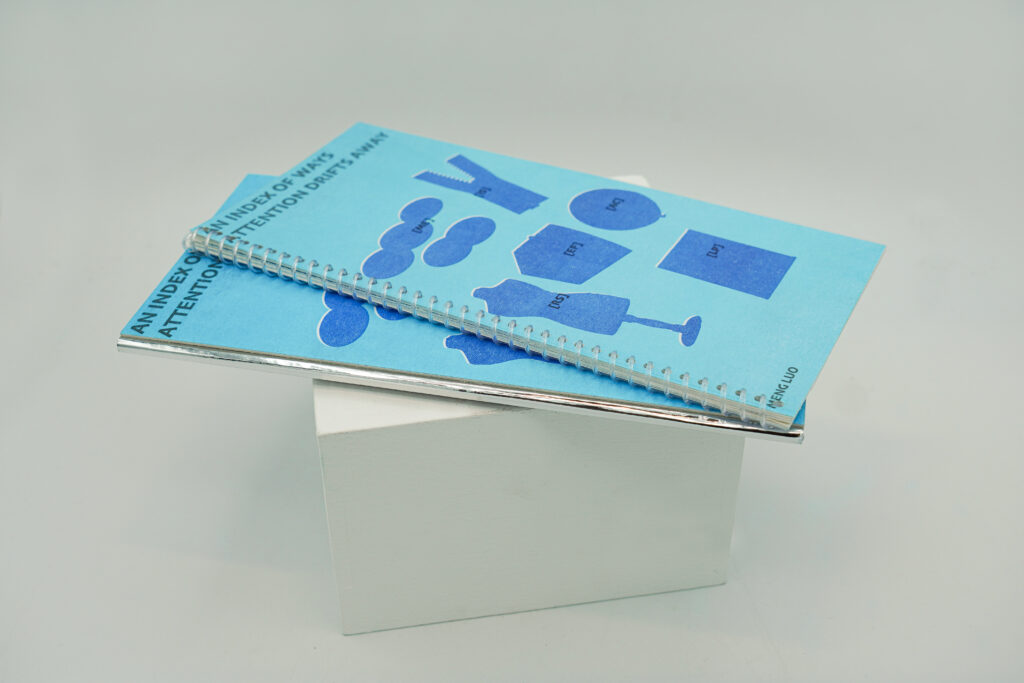 Two physical editions of An Index of Ways Attention Drifts Away are displayed together on a white block, showing the front covers of both publications.