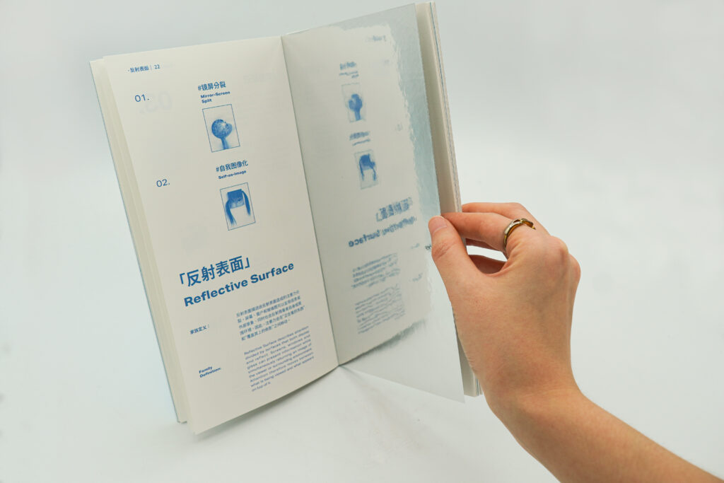 A public publication showcasing a series of reflective mirror-like surfaces. One hand flipped over the reflective paper on the right page, on which the blurry blue image and text on the left page were reflected.