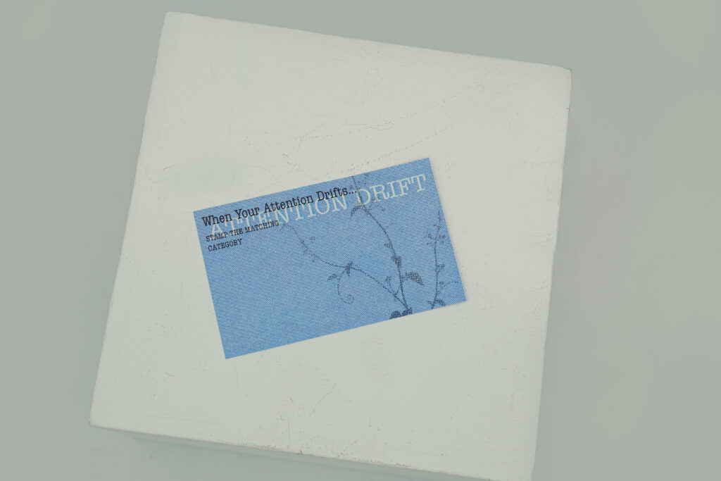 A single blue Attention Drift Observation Card rests on a white display block. The card contains printed instructions reading When Your Attention Drifts and a faint vine graphic in the background.
