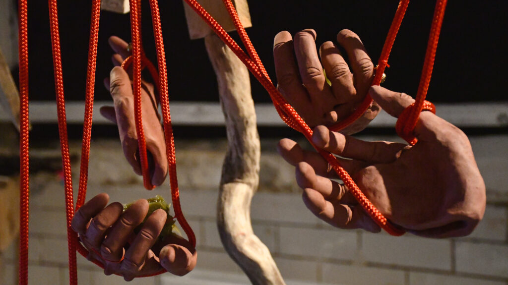 hands made of red clay tangled in red rope and suspended from a wooden structure hanging in the air, some of the hands hold blobs of green ointment