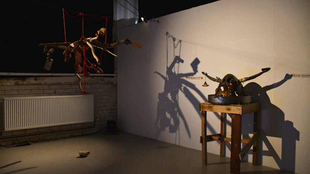 a soft spotlight in a dark space illuminates a hanging sculpture, casting a branching shadow onto the wall and over a large ceramic head with outstretched arms that sits on a wooden plinth.