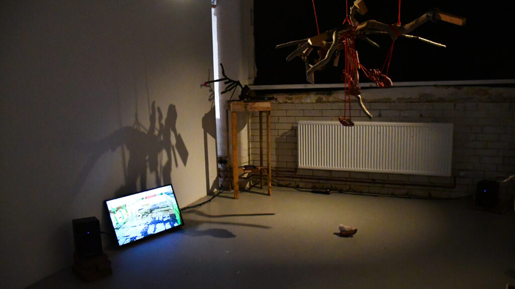 a dark space lit by a soft spotlight with a speaker and tv on the floor next to a wooden plinth on which a burned-wood bird sculpture with outstretched wings rests. In the middle of the space is a suspended sculpture with rope hanging from the centre