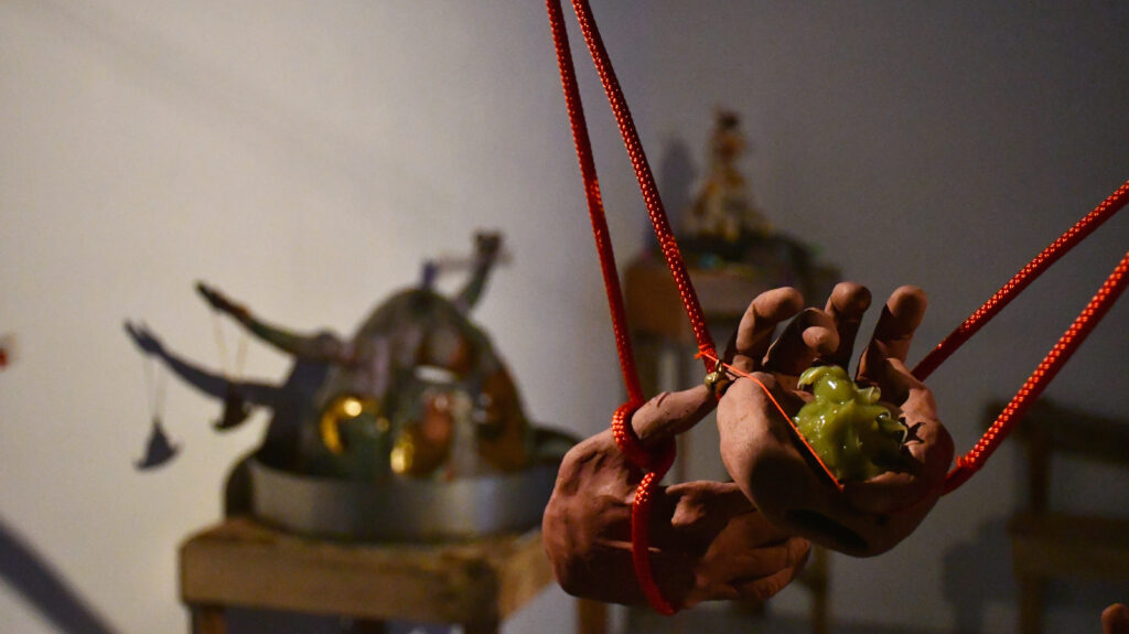 two red clay hands suspended in the air by red rope and thread twined around the fingers, one thread has a small brass bell, one hand holds a large blob of green ointment. In the background, out of focus, a large blue/grey/purple/red conical ceramic head with outstretched arms sits in a metal dish on a wooden plinth and holds a triangular gong. Further back a pale horse puppet sits on a grey cushion on top of a tall wooden plinth.