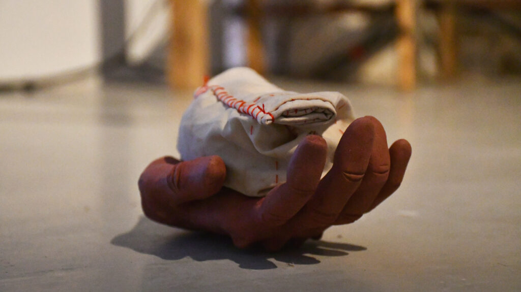 a red clay hand rests on the floor with the palm upturned. It is holding a white cotton parcel stitched with red thread.