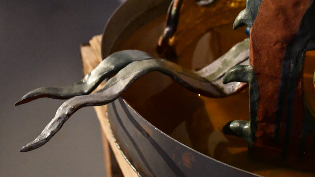 a ceramic forked tongue glazed metallic blue and purple ripples out from a carved mouth with triangular teeth along the sides, the tongue reaches out over the rim of a metal bowl