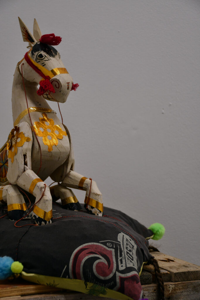 a white-painted wooden horse puppet with gold detailing sits on a grey cotton and gold silk cushion with red stitching, there are neon green, neon yellow, and bright blue pom poms