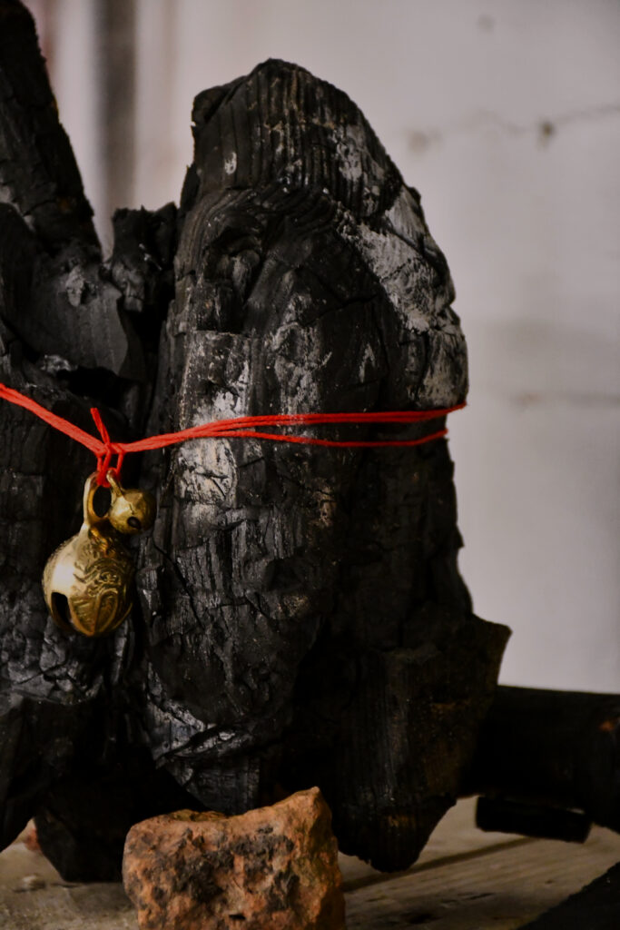 a black burnt piece of wood with white ashy patches stands supported by a small piece of orange brick, around the charcoal lump is two wraps of red thread with two brass bells of different sizes hanging at the front