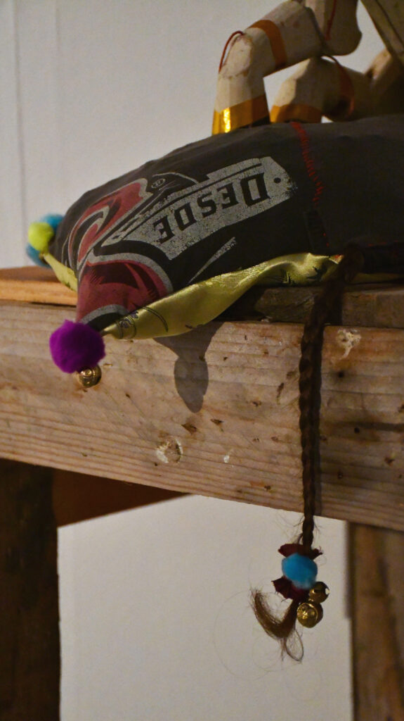a grey cushion with purple pom pom on one corner, and neon yellow pom pom on another corner, a plait of hair finishing with a blue pom pom and bell overhangs the wooden plinth on which the cushion sits