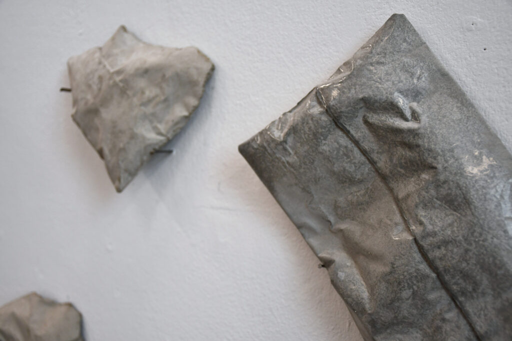 three grey concrete forms, one shiny and rectangular, the others dull, smaller, and irregular in shape arranged in a triangular shape and balanced on nails hammered into a white wall