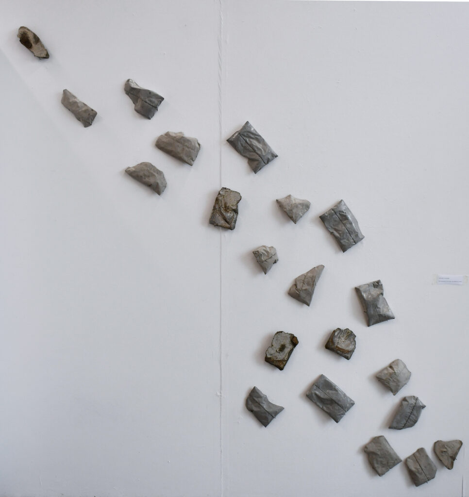 21 grey concrete forms, all of similar size and most of them rectangular, are arranged in a flowing shape, like a shoal of fish, that travels up a white wall