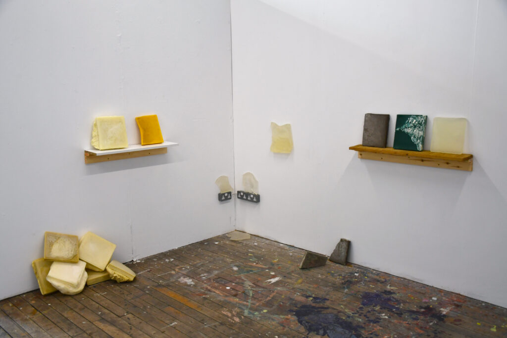 in the corner of a white room with wooden floor, one shelf on the left wall holds two cast books, a shelf on the right wall holds three books of different colours, a further book is balanced on the wall in between with broken fragments and discarded books littering the floor and in a pile