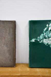 a grey concrete cast of a book stands on a wooden shelf next to a green wax cast of the same book, Burma to Myanmar is embossed in the top left corner of the green wax cast