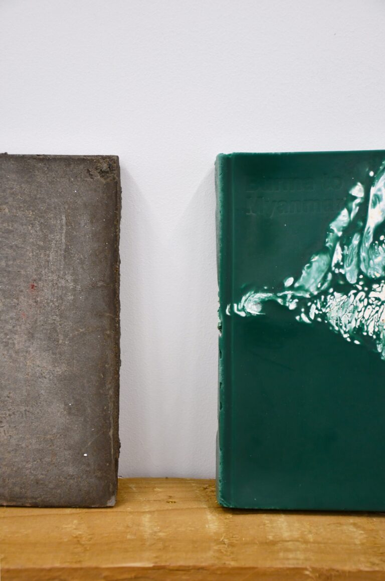 a grey concrete cast of a book stands on a wooden shelf next to a green wax cast of the same book, Burma to Myanmar is embossed in the top left corner of the green wax cast