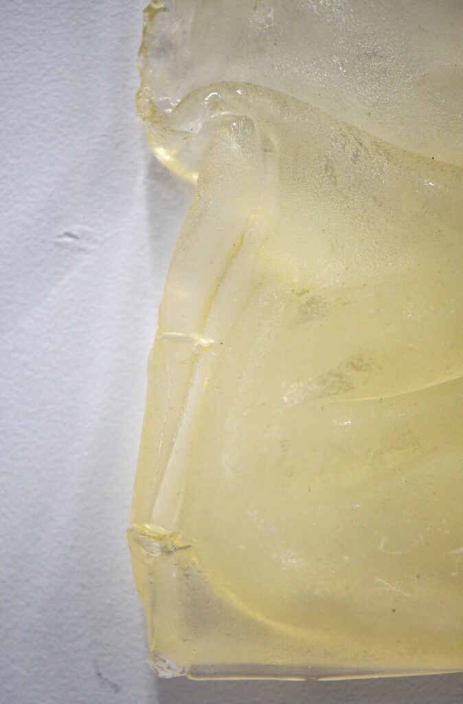 a clear rectangular form slumps and folds as it leans against a white-painted wall